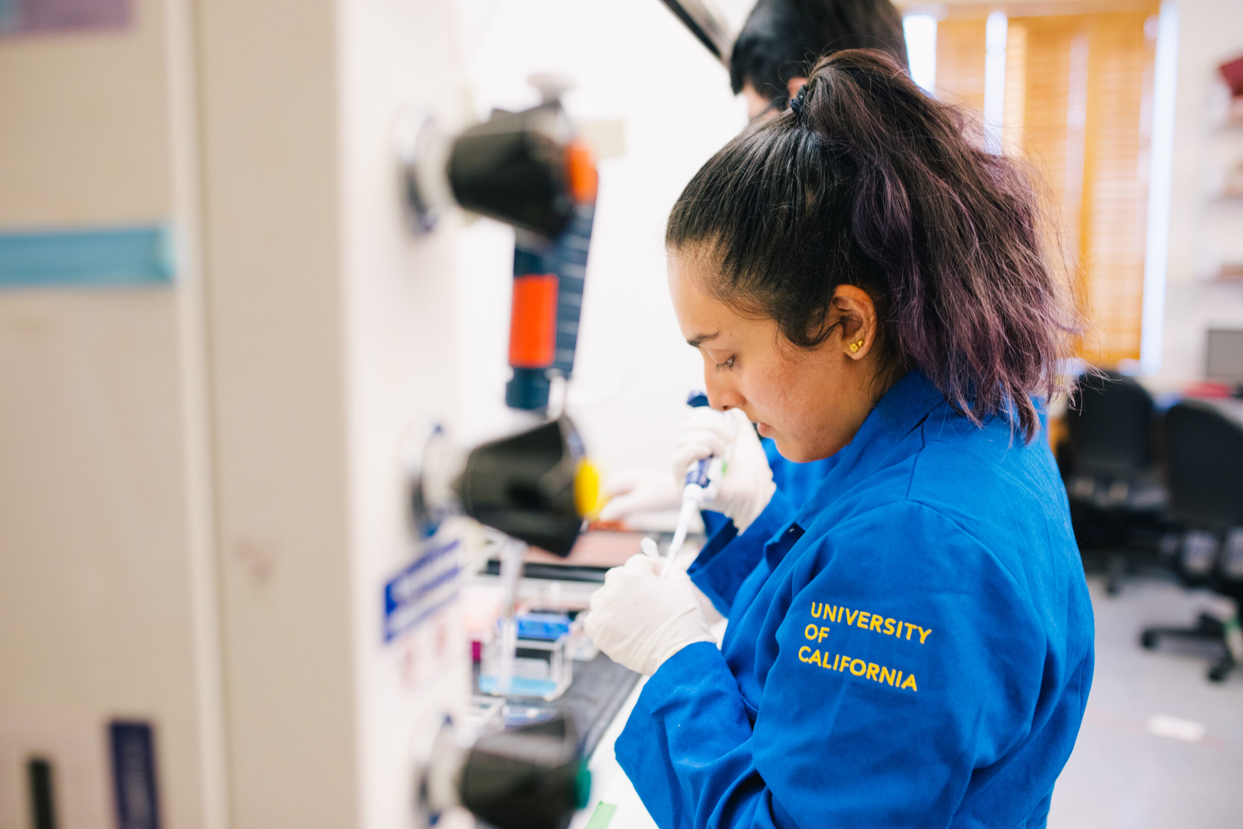 A scientist wearing a blue lab coat that reads University of California on the left arm stands holding a pipette before a machine.