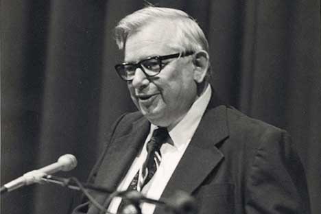 Older man with white hair, horned rim glasses speaking into a microphone