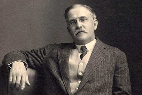 Man wearing pin-striped suit and glasses with arm resting on chair