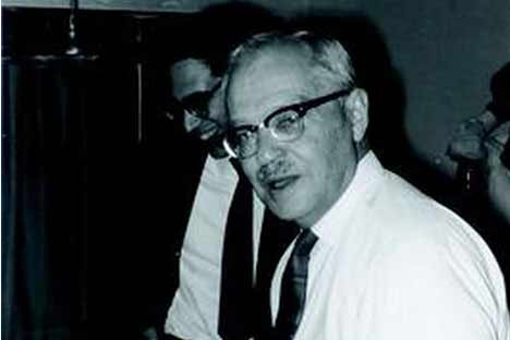 Black and white photo of man wearing horned-rim glasses, tie, and looking at viewer
