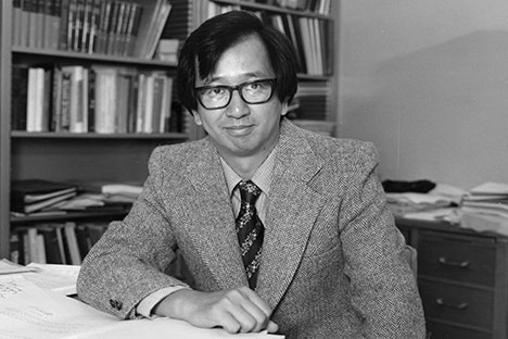 Man with black hair wearing horn-rimmed glasses, suit and tie sitting with arm resting on desk