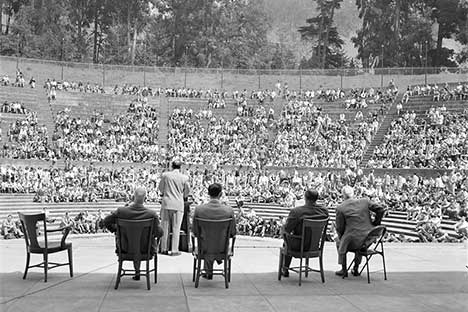 View from the rear of a man speaking to students seated in Greek Theatre. Four men seated behind the speaker.
