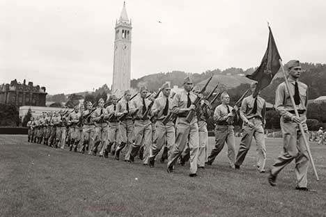 Black and white photo of marching uniformed servicemen with Campanile in background