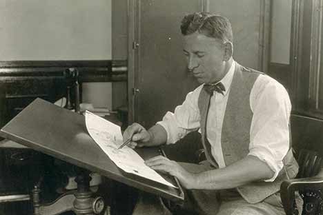 Black and white photo of man wearing vest and bowtie drawing at a tilted table