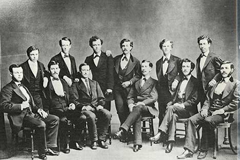 Historic black and white photo of 12 men wearing suits and bowties sitting and standing