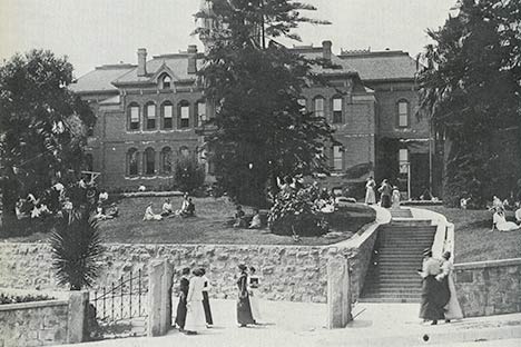 Black and white photo of the facade of a building with stairs leading to it and people walking and sitting in front