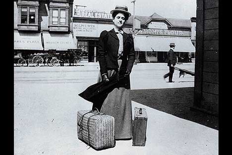Black and white photo of woman wearing with two suitcases. Horsedrawn cart and man pushing wheelbarrow in background.