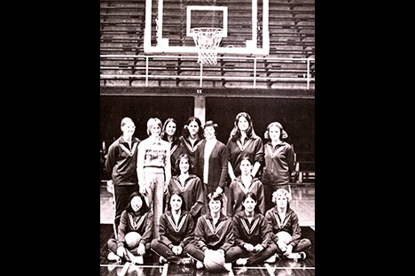Group of women sitting and standing below a the basket on a basketball court.